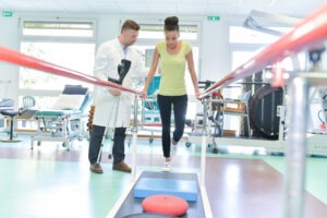 A woman in a yellow shirt holding onto rails is next to a physical therapist in a white lab coat.